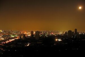 "A cityscape at night illuminated by scattered lights and the full moon, with high-rise buildings and glowing streets under a hazy, orange-tinged sky."