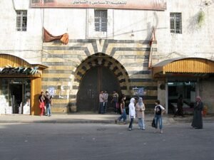 A group of people stands in front of a large, arched stone gate with black and yellow stripes, part of an old building with Arabic text on a sign above. The scene is on a street with small shops on either side, one with a wooden awning and the other displaying goods outside. People are casually walking or standing, some in traditional attire, with a few flags visible near the shops.