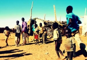 A group of children and adults stands on a dusty ground in a rural area under a clear blue sky. They are dressed in casual, worn clothing, with some children barefoot. In the background, there are makeshift structures made of wood and mud, with sticks protruding from the roofs. One child in a blue shirt is captured mid-run, casting a shadow on the ground.